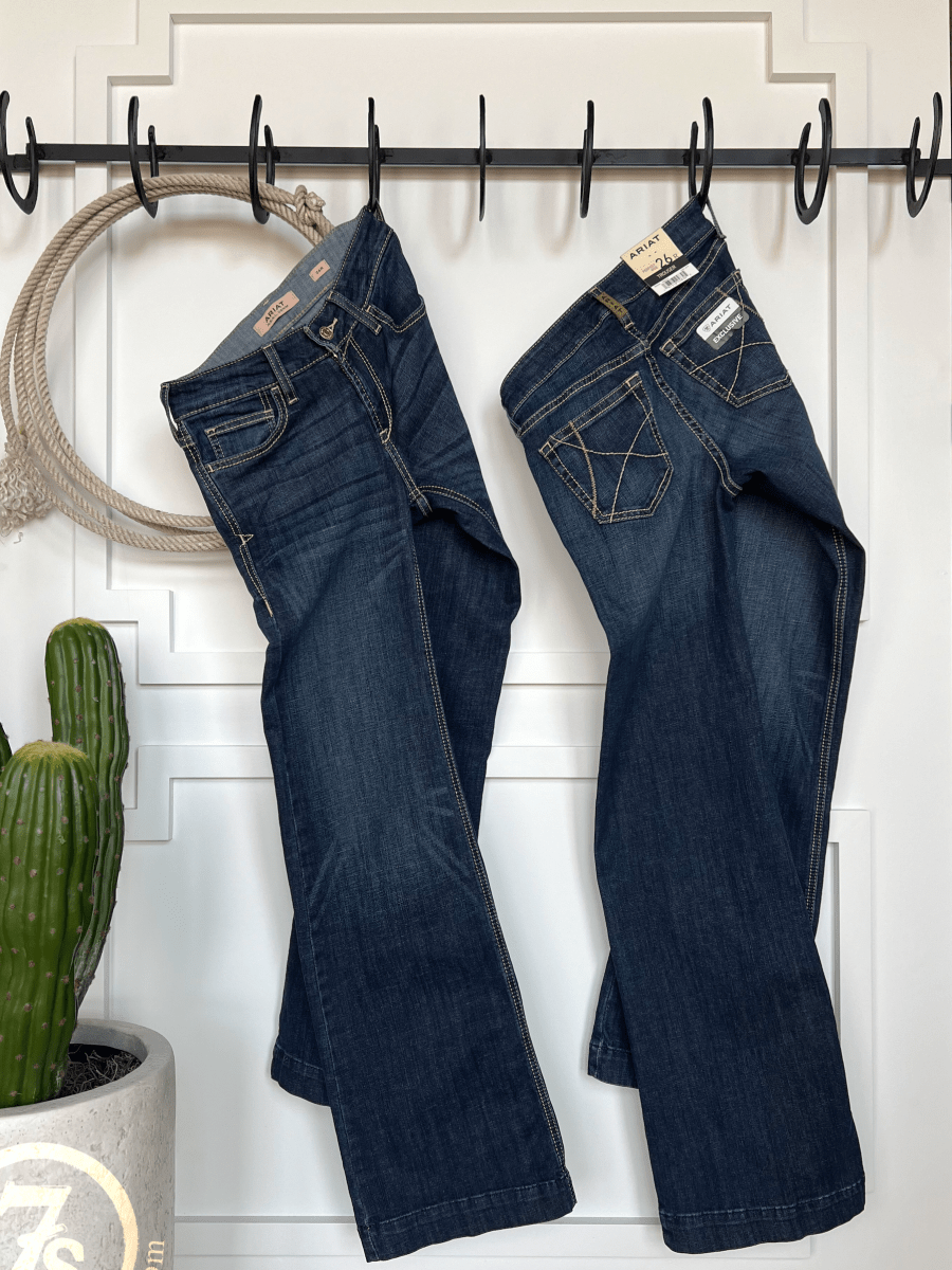 Two pairs of The Blayze Perfect Rise Trouser Jean by Savannah Sevens western life{&}style hang on black hooks against a white paneled wall, with a potted cactus and coiled rope accenting the comfy, no-gap design.