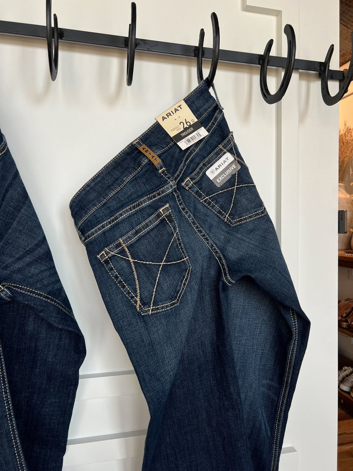 A pair of The Blayze Perfect Rise Trouser Jeans by Savannah Sevens western life{&}style in medium dark wash with white contrast stitching hang from a black metal hook rack on a white paneled wall. Tags show size 26 regular and a comfy no-gap waist.