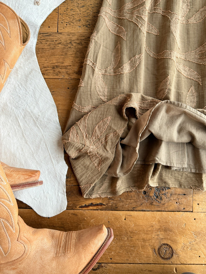 Tan cowboy boots beside the Bluff City Sage Floral Dress by Savannah Sevens western life{&}style, featuring petite adjustable straps and a ruffled hem, displayed on a rustic wooden floor overlapping a light cowhide rug.