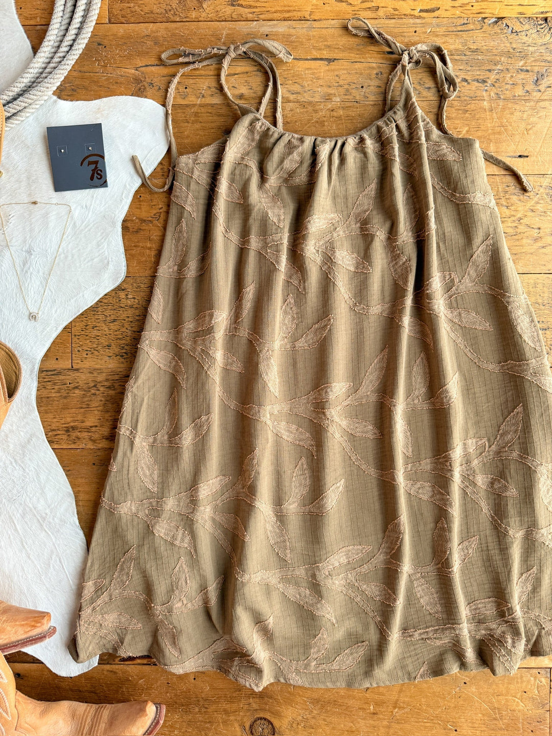 The Bluff City Sage Floral Dress by Savannah Sevens western life{&}style, a light brown sleeveless dress with petite adjustable straps and an embroidered bird pattern, is laid flat on wood beside a cowboy boot, necklace, tag 78, and coiled rope.