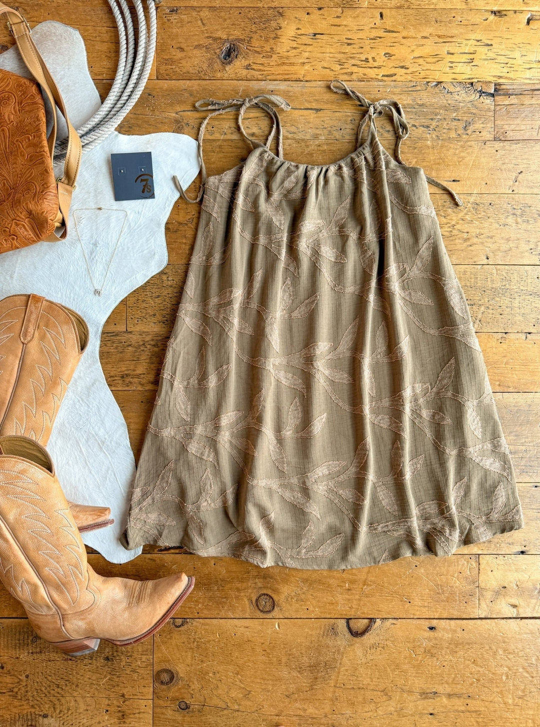 The Bluff City Sage Floral Dress by Savannah Sevens western life{&}style, with petite adjustable straps, is styled on a wooden floor with tan cowboy boots, a brown leather bag, jewelry, and rope for a rustic Western-themed flat lay.
