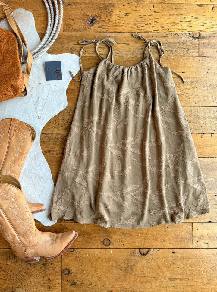 The Bluff City Sage Floral Dress by Savannah Sevens western life{&}style, with petite adjustable straps, is styled on a wooden floor with tan cowboy boots, a brown leather bag, jewelry, and rope for a rustic Western-themed flat lay.