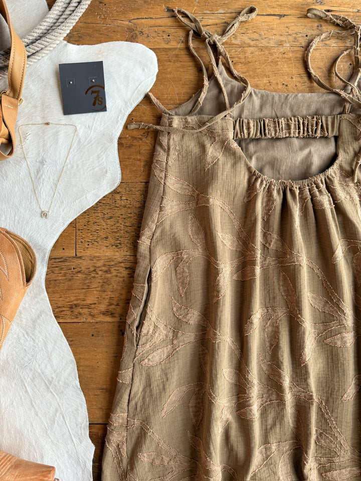 The Bluff City Sage Floral Dress by Savannah Sevens western life{&}style, a brown sleeveless style with petite adjustable straps, is displayed on wood beside tan sandals, a delicate necklace, earrings on a card, and white textured fabric.