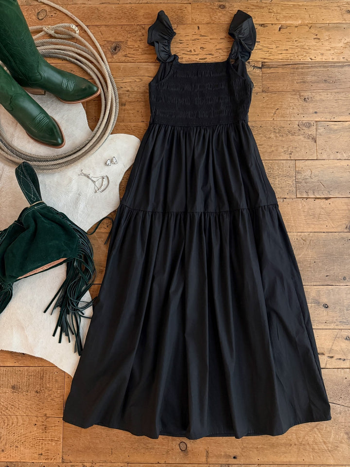 The Boca Raton Black Dress by Savannah Sevens western life{&}style is styled on a wooden floor with green cowboy boots, silver jewelry, a green fringed purse, and a white cowhide rug.