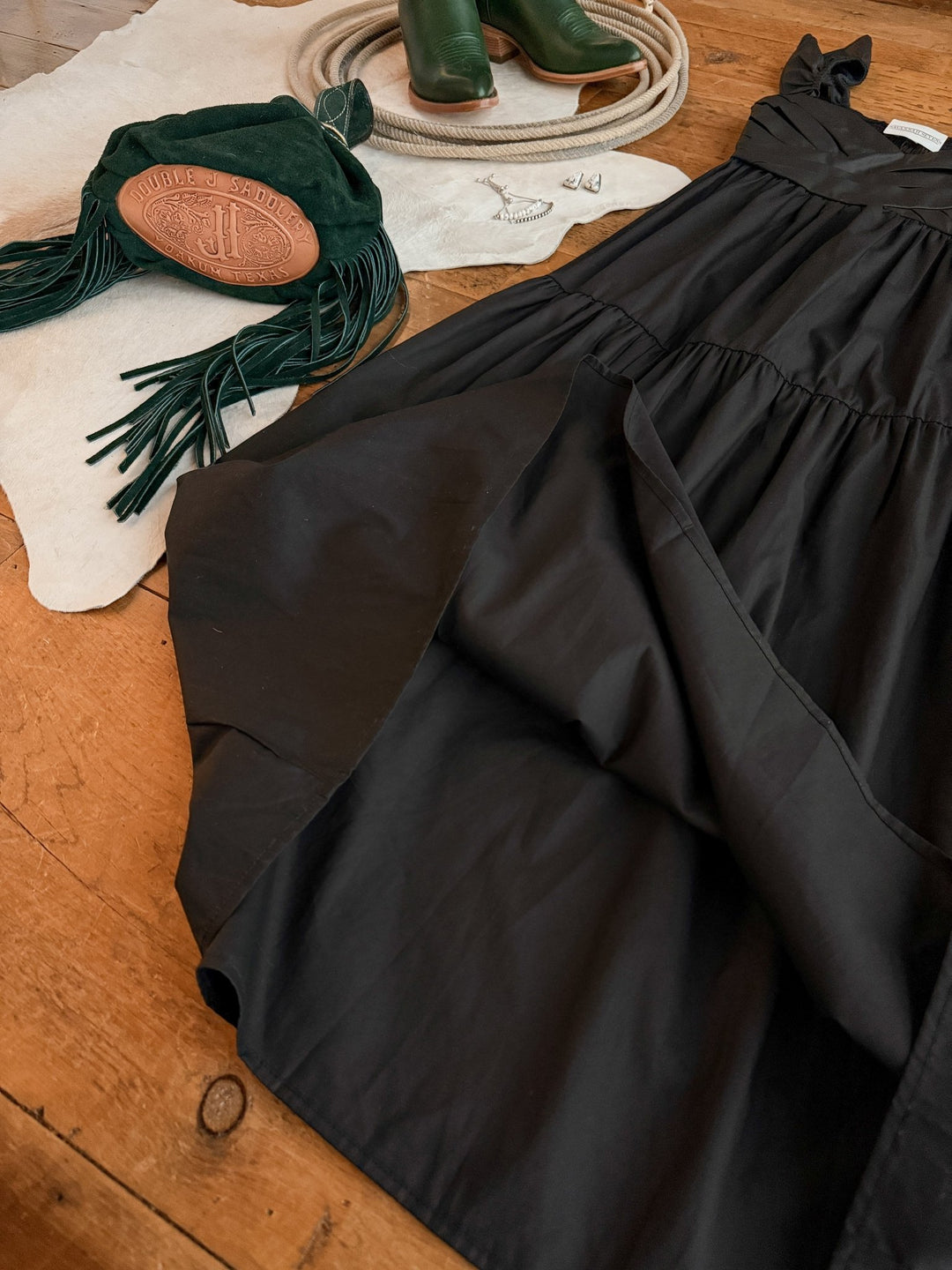 The Boca Raton Black Dress from Savannah Sevens western life{&}style rests on wood beside a green fringe bag, tan leather clutch, green boots, and silver jewelry atop cream fabric.