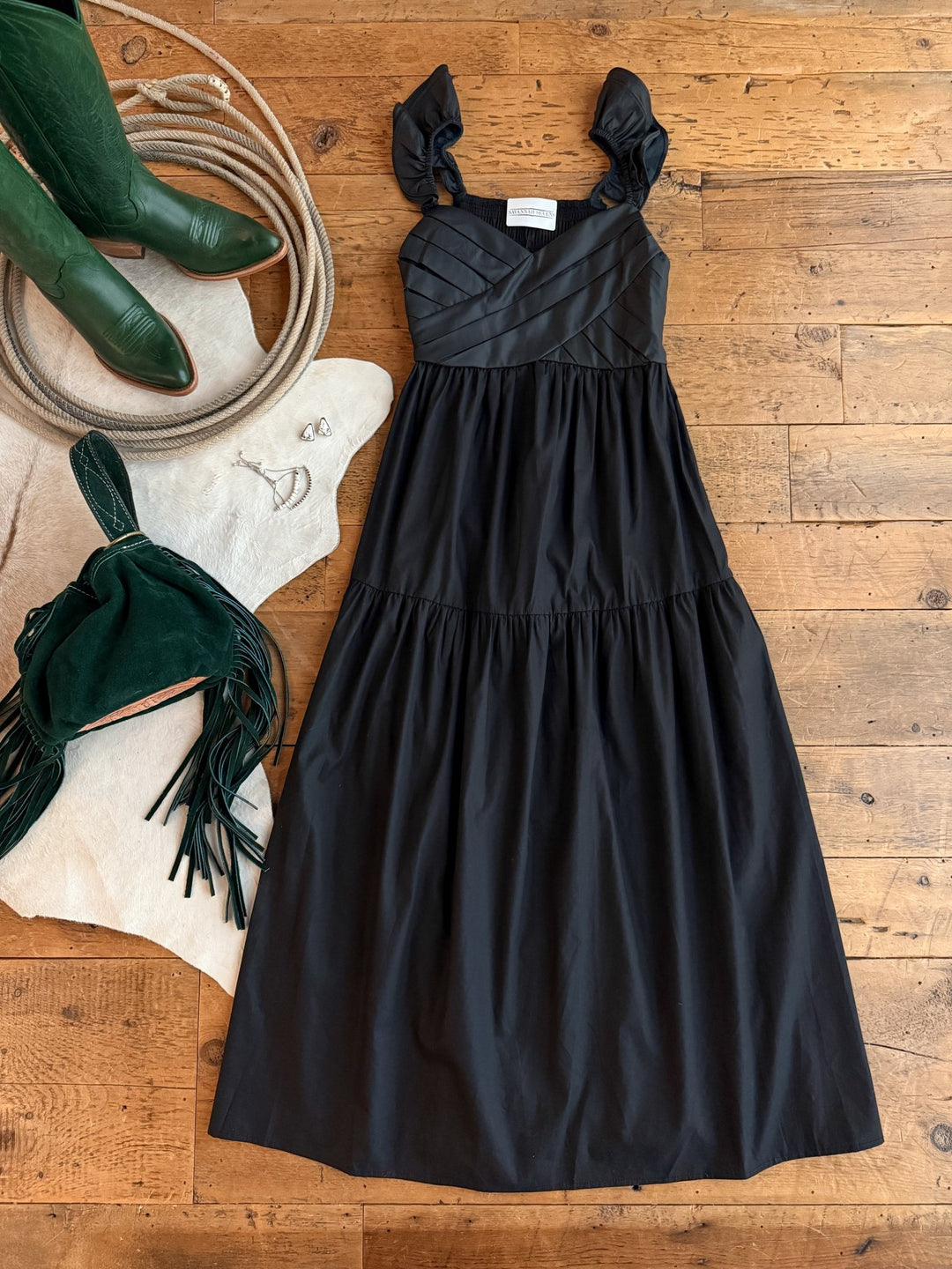 The Boca Raton Black Dress by Savannah Sevens western life{&}style is laid out on a wooden floor with green cowboy boots, silver jewelry, and a green fringed bag atop a white cowhide rug.