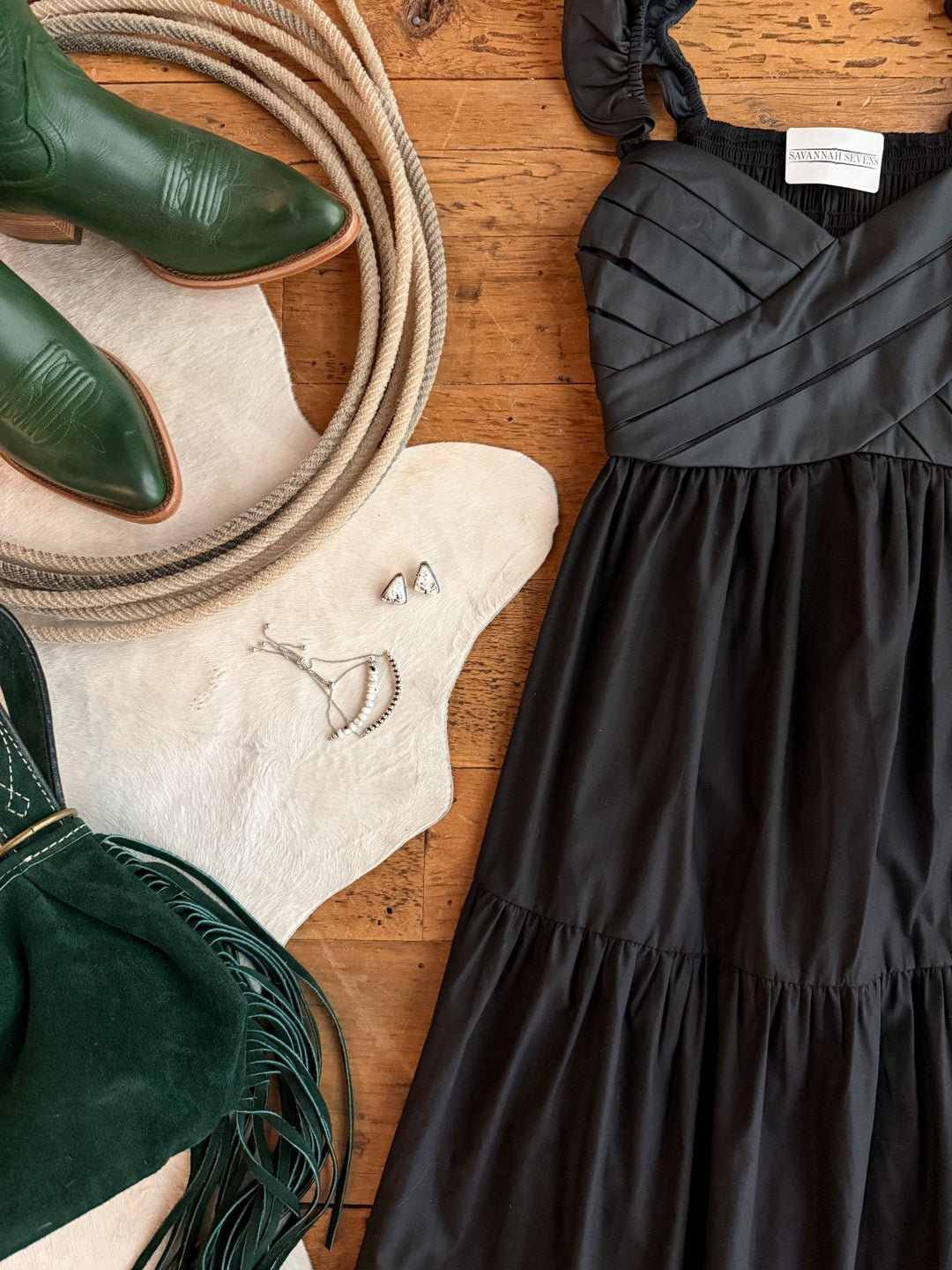 The Boca Raton Black Dress by Savannah Sevens western life&style, green cowboy boots, a coiled rope, green suede fringe purse, silver jewelry, and a white cowhide are displayed on a wooden floor.