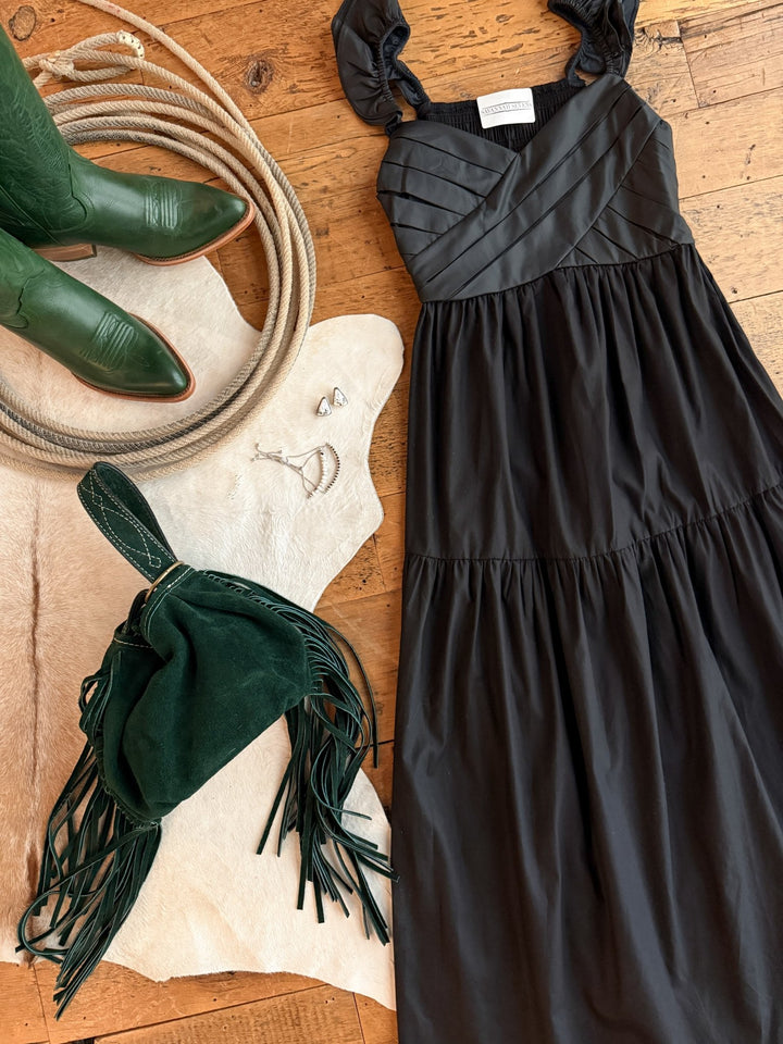 The Boca Raton Black Dress by Savannah Sevens western life{&}style, paired with green cowboy boots, a green fringed bag, a white cowhide rug, and silver jewelry, is arranged on a wooden floor.