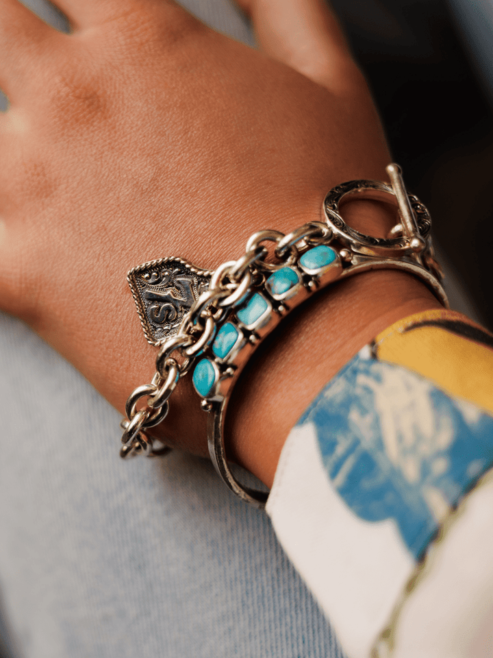 A close-up of a hand wearing the Boca Raton Bracelet by Savannah Sevens western life{&}style, featuring turquoise stones and an ornate charm, resting on light blue denim with part of a colorful, patterned sleeve visible.