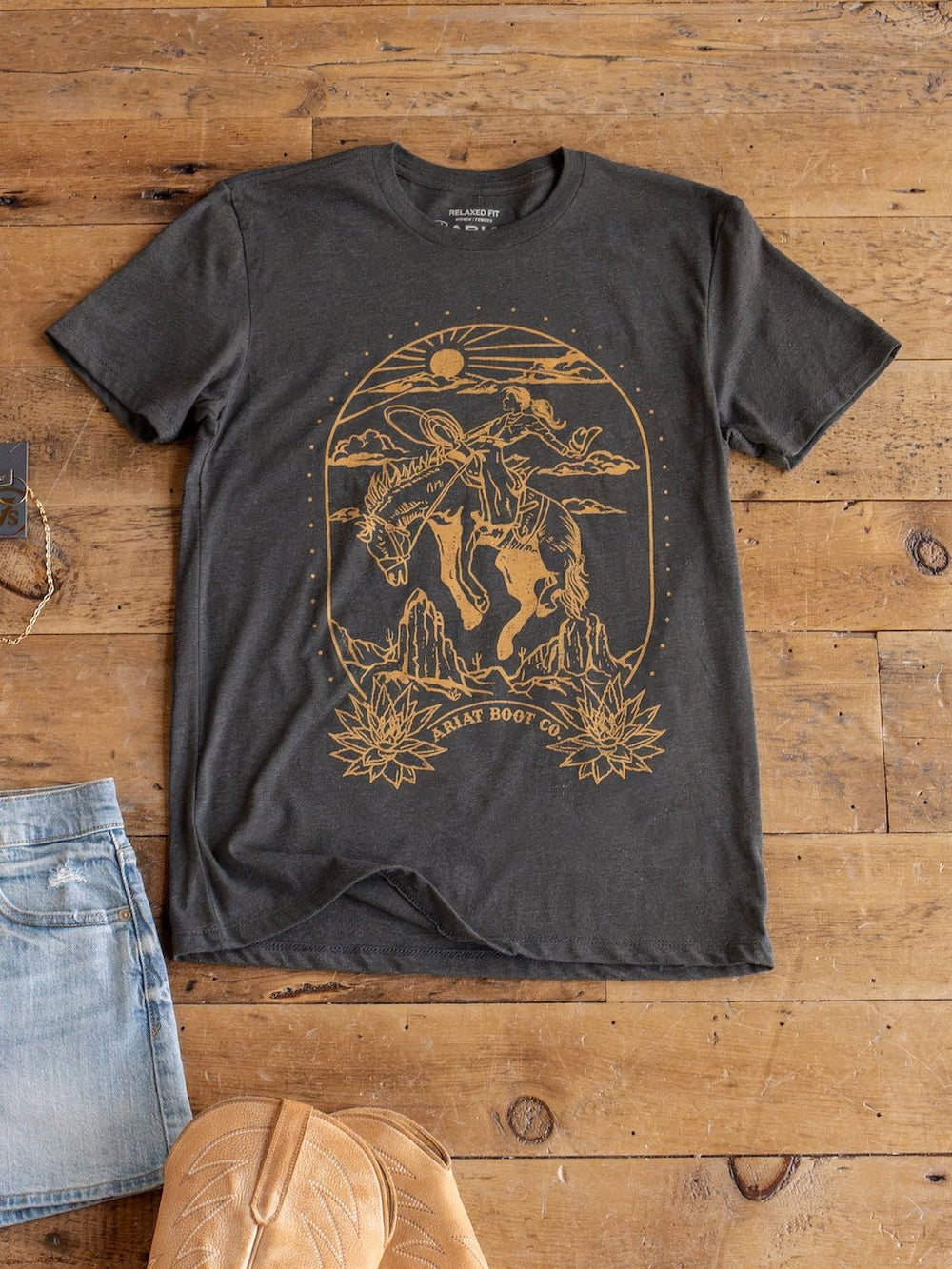 A Boot Co. Charcoal Western Graphic Tee from Savannah Sevens western life{&}style, featuring a yellow print of a cowboy riding a horse under the sun and surrounded by cacti, with "Ariat Boot Co." at the bottom. This unisex fit shirt is displayed on a wooden floor alongside denim shorts and tan boots.