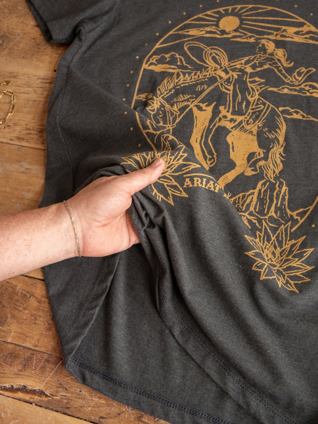 A hand pinches the Boot Co. Charcoal Western Graphic Tee by Savannah Sevens western life{&}style, featuring a gold cowboy on horseback, desert plants, and a sun graphic, all laid out on a rustic wooden surface.