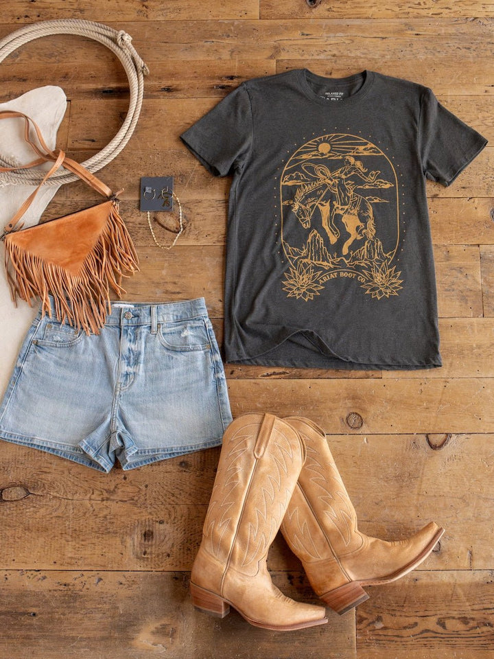 Boot Co. Charcoal Western Graphic Tee by Savannah Sevens western life{&}style, cowboy boots, denim shorts, a fringe bag, and jewelry are arranged on a wooden floor in a flat lay, showcasing a unisex western-inspired outfit.