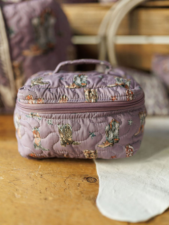 A quilted purple fabric bag from the Boots Collection by Savannah Sevens western life&style, featuring whimsical cat and star motifs, a zipper, and handle, rests on a wooden surface with similar bags softly blurred in the background.