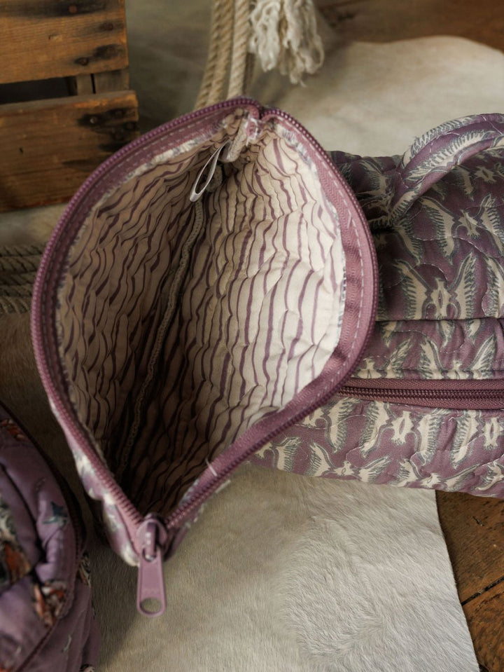 A purple-patterned fabric pouch with a quilted light interior from Savannah Sevens western life{&}style’s Boots {Collection} rests on a surface, surrounded by other pouches and a wooden crate in the background.