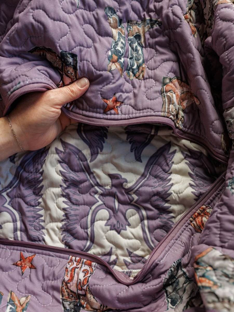 A hand opens a zipped section of the purple quilted Boots {Collection} bag by Savannah Sevens western life{&}style, revealing an inner layer with a bold eagle graphic on a cream background.