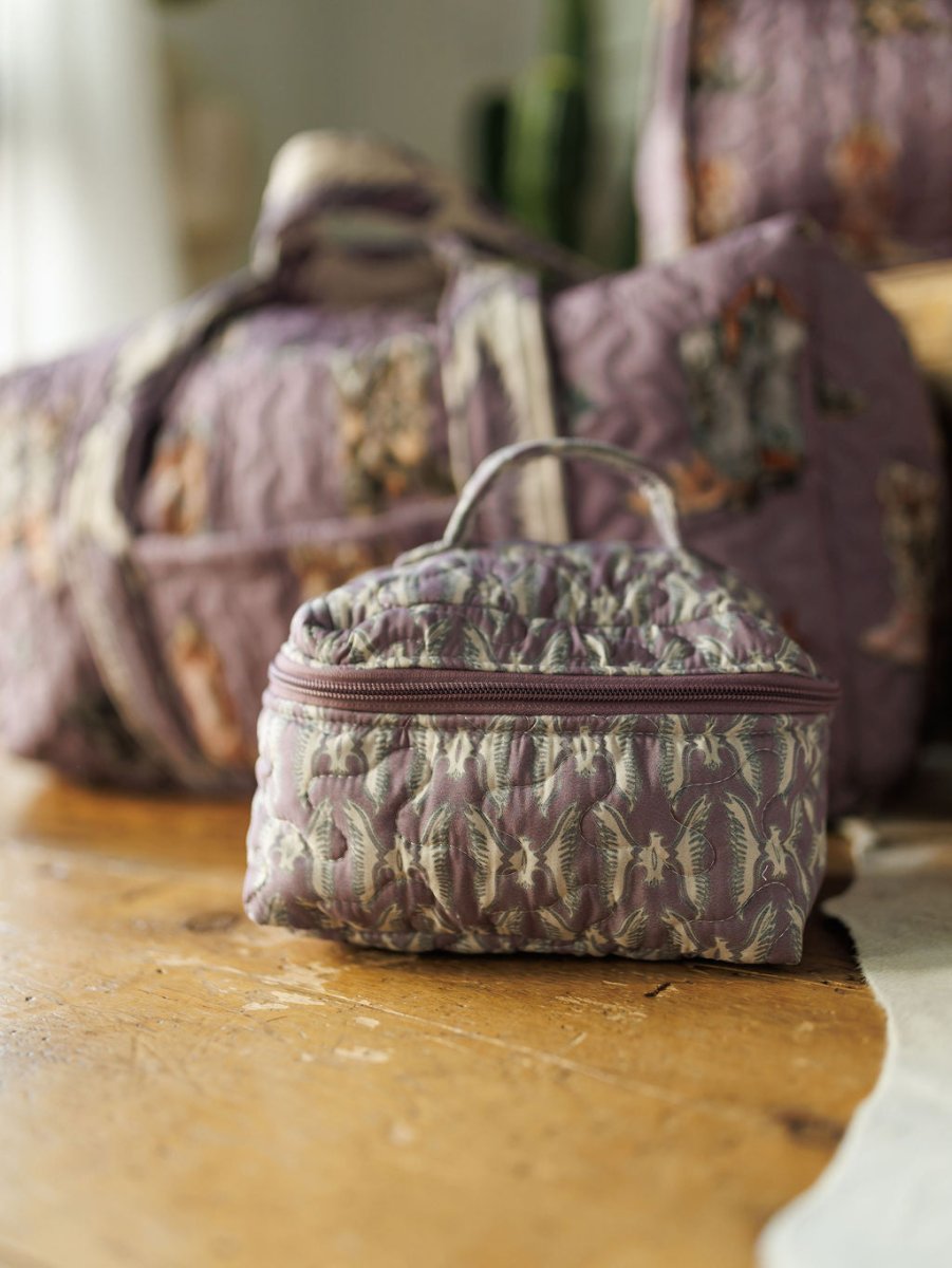 A small, quilted purple floral travel bag with a zipper from the Boots Collection by Savannah Sevens western life&style rests on a wooden surface, with a matching larger quilted bag blurred in the background.