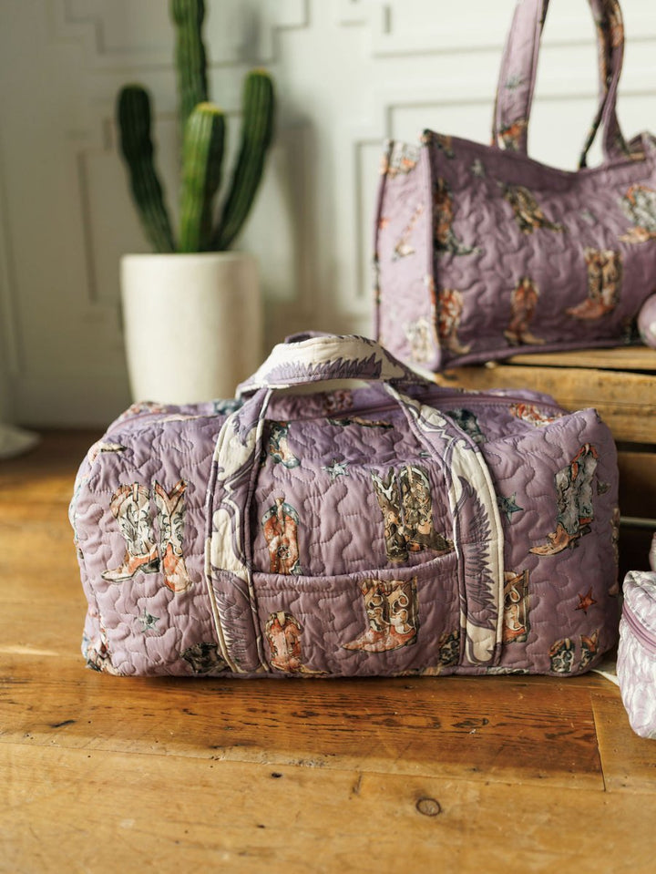 A quilted Boots Collection bag set in lavender with playful animal prints by Savannah Sevens western life{&}style rests on a wooden floor. A matching bag and potted cactus sit nearby against a white paneled wall, evoking handmade charm.