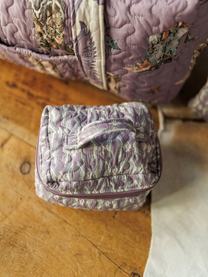 A small, handmade quilted purple bag with a handle from the Boots Collection by Savannah Sevens western life{&}style sits on a wooden floor beside a matching floral-patterned item, both part of the same quilted bag set.