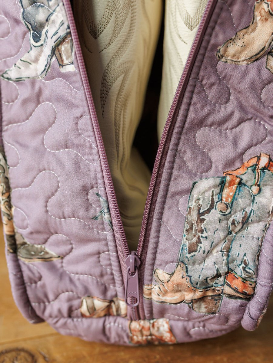 Close-up of a quilted bag from the Boots Collection by Savannah Sevens western life{&}style, featuring cowboy boots and hats on a purple background with a purple zipper. The bag is partially open, showing cream-colored contents inside.