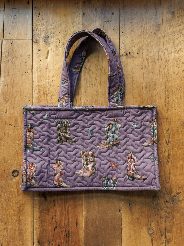 A rectangular purple handmade quilt tote from the Boots Collection by Savannah Sevens western life{&}style, featuring a cowboy boot pattern and two handles, displayed on a rustic wooden floor. Part of the western travel collection.