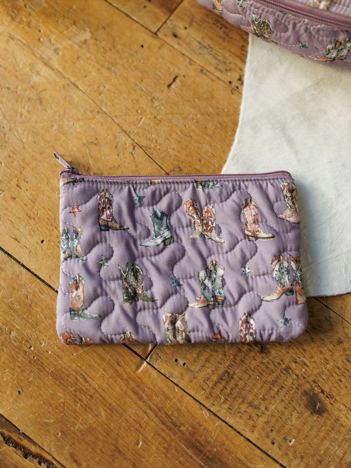 A small quilted lavender pouch from the Boots Collection by Savannah Sevens western life&style, featuring a colorful cowboy boots pattern and zipper, rests on a wooden surface beside white fabric.