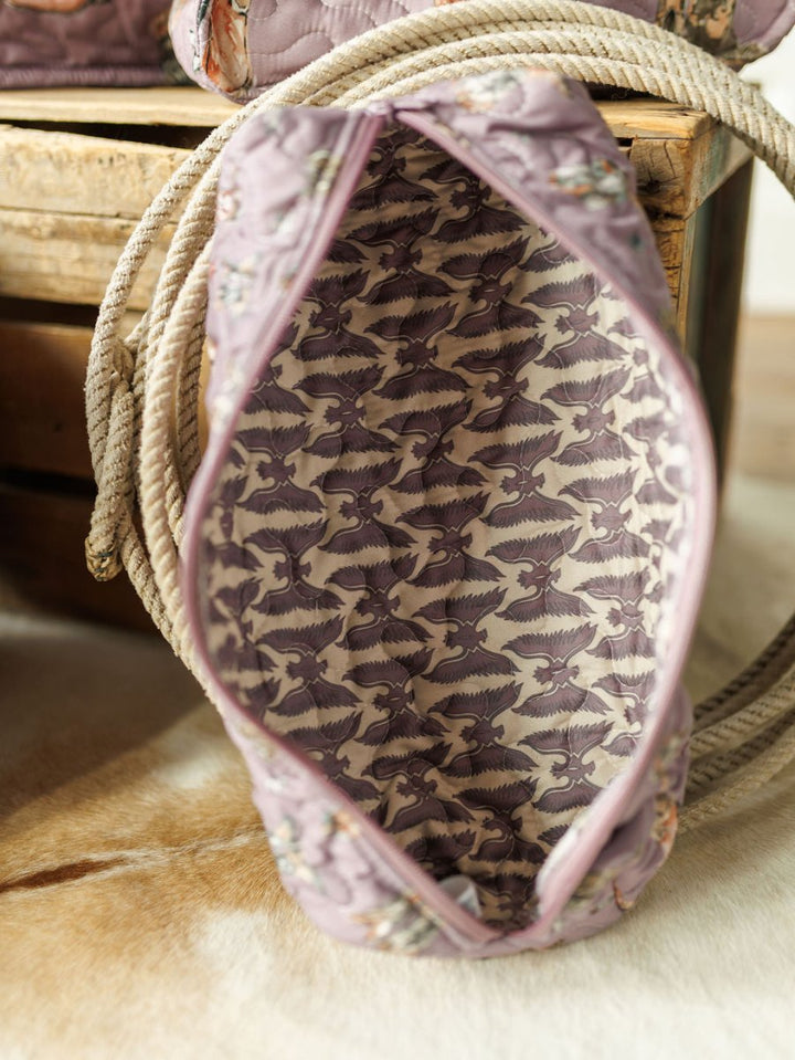 A close-up of a handmade quilt bag from the Boots Collection by Savannah Sevens western life{&}style, lined with light purple fabric featuring white cranes, resting on a wooden crate beside a coiled rope.