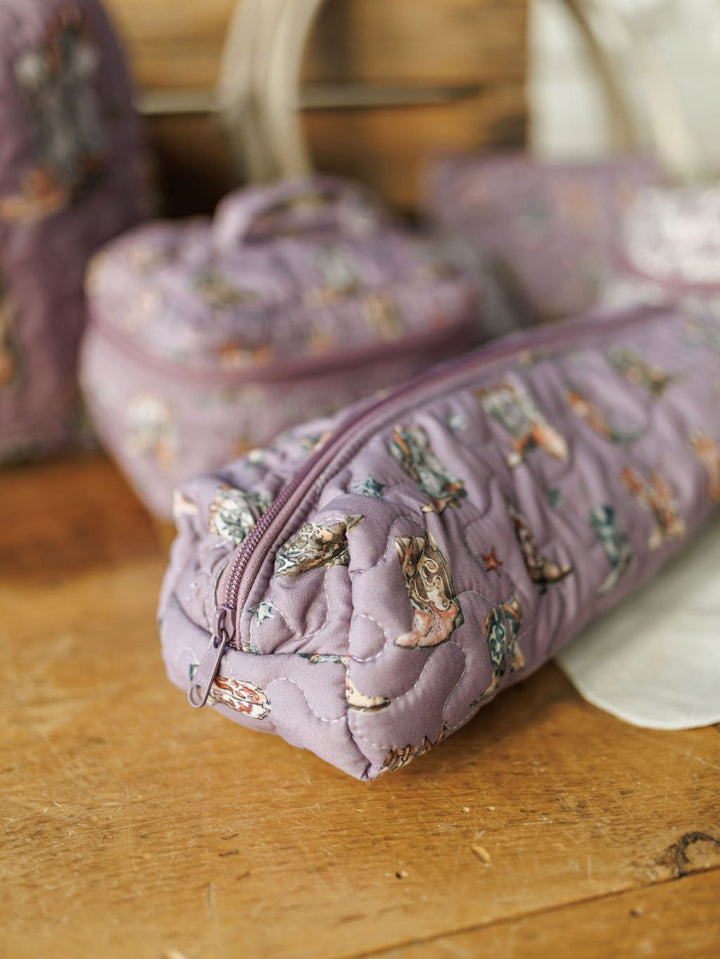 A quilted lavender pencil case with whimsical animal patterns from the Boots Collection by Savannah Sevens western life{&}style rests on a wooden surface, with matching quilted bags in the background.