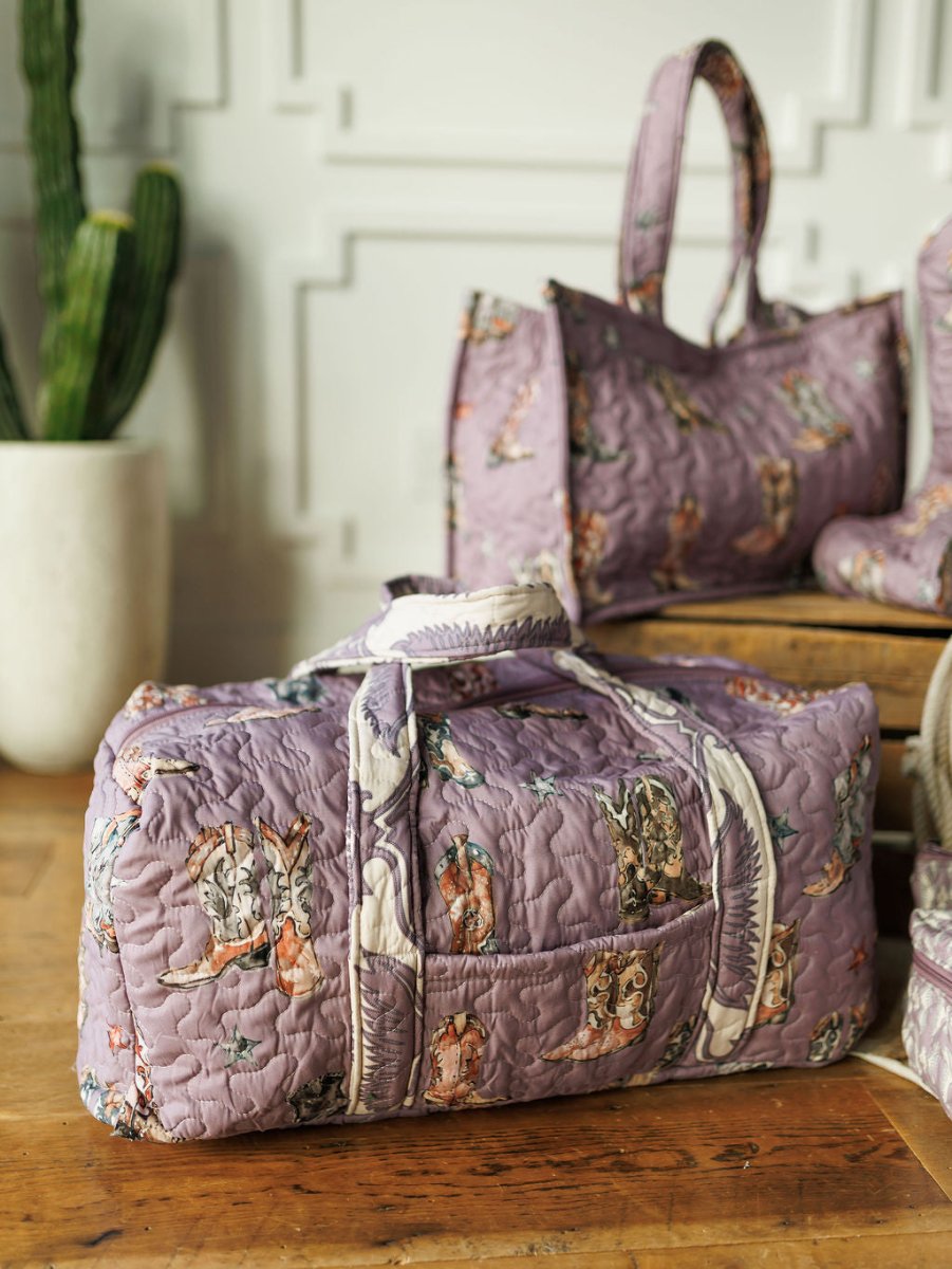 A lavender quilted duffel from the Boots Collection by Savannah Sevens western life{&}style, featuring animal prints, sits on a wooden surface with matching bags and a potted cactus against a white textured wall.