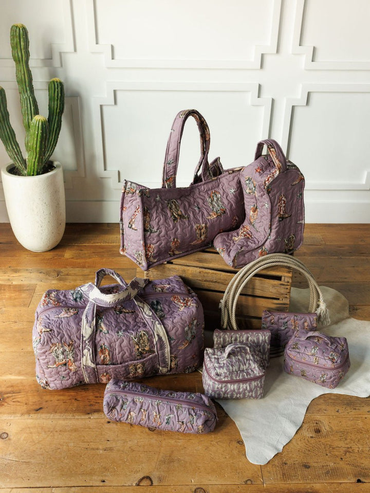 The Boots {Collection} by Savannah Sevens western life{&}style features purple quilted bag sets with embroidered animals, displayed on a wooden floor beside a cactus and white wall, highlighting this handmade quilted western travel set.
