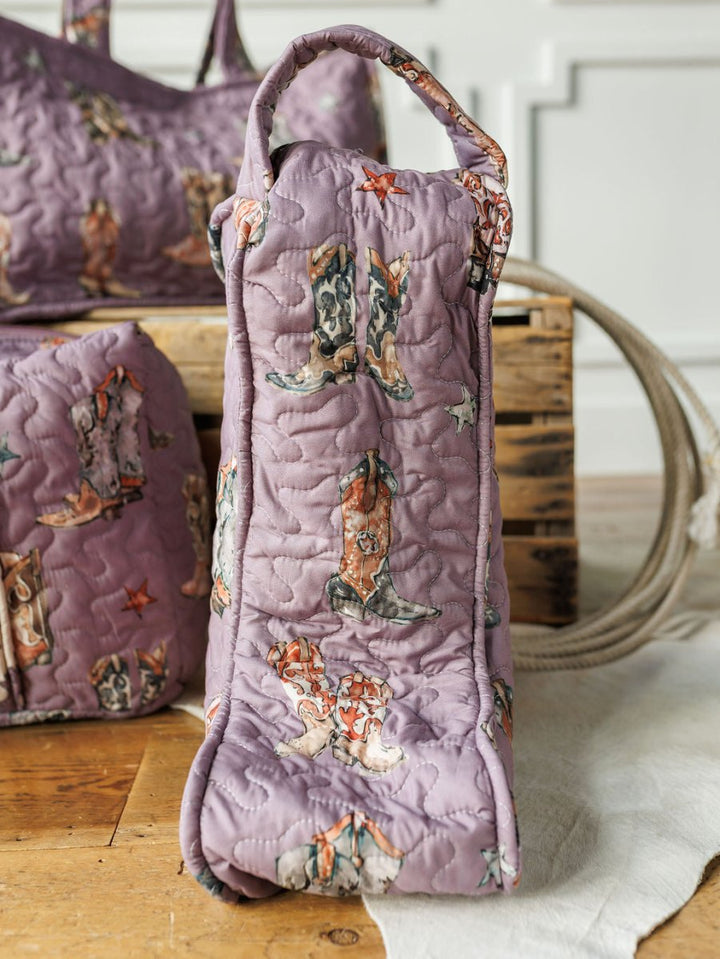 A handmade quilted lavender bag from the Boots {Collection} by Savannah Sevens western life{&}style stands upright on a wooden floor, with a matching bag, crate, rope, and white paneled wall in the background.