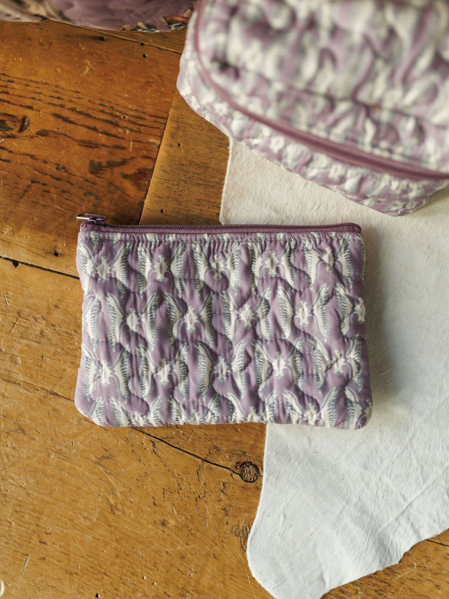 A small purple quilted pouch from the Boots {Collection} by Savannah Sevens western life{&}style, featuring a zipper, rests on a wooden surface near a white cloth. Another quilted item from the collection is partially visible in the corner.