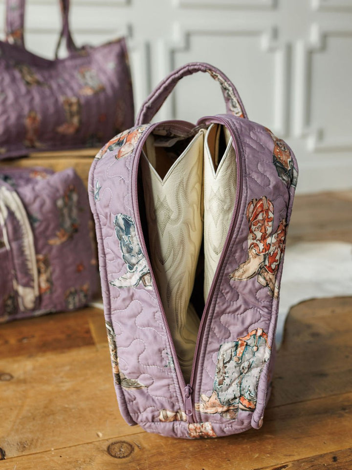 A purple floral quilted bag from Savannah Sevens western life{&}style's Boots Collection is unzipped to reveal white cowboy boots inside, resting on wood, with another matching bag from the collection in the background.