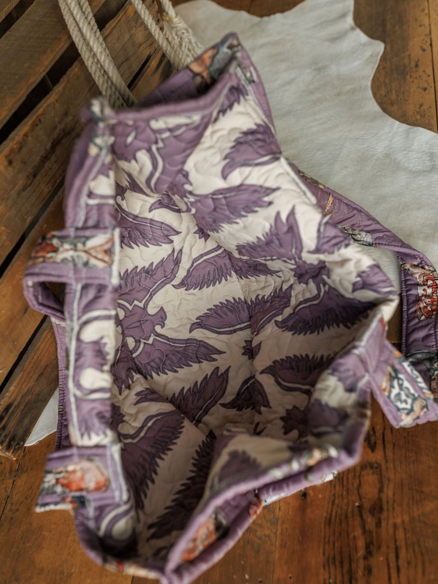 The Boots Collection tote by Savannah Sevens western life&style features purple and cream leaf-patterned lining, rope handles, and a spacious interior. Shown on wood beside a white cowhide rug.