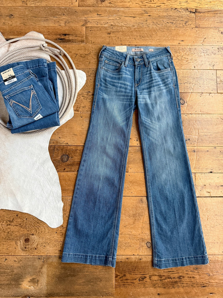 A pair of The Bowen Perfect Rise Trouser Jeans by Savannah Sevens western life{&}style is laid flat on a wooden floor beside folded medium wash jeans and a coiled rope on a white cowhide rug.