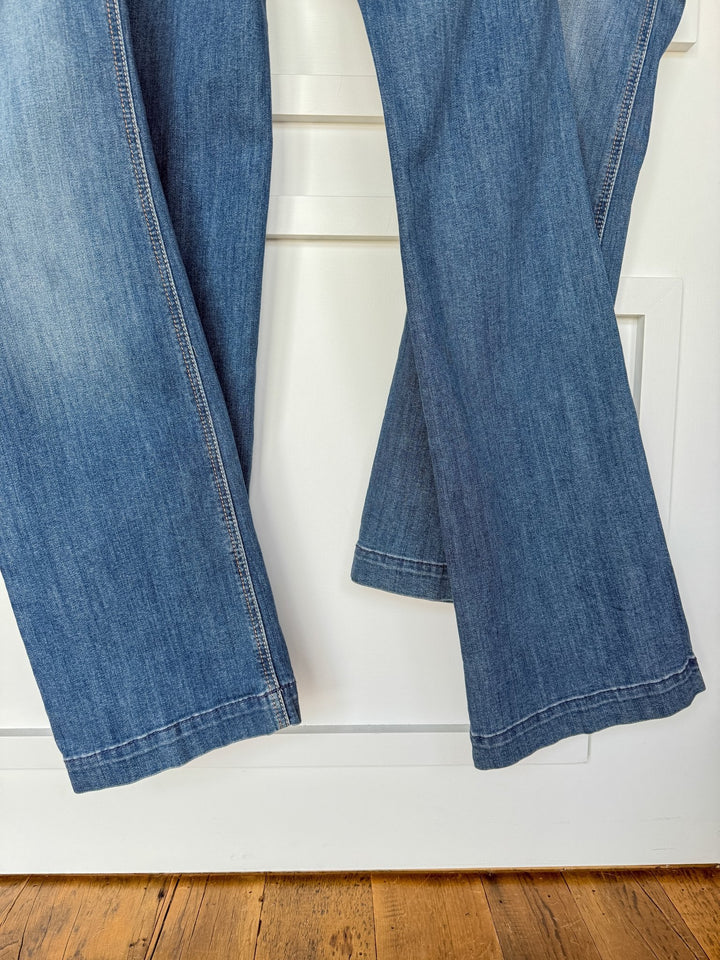 A close-up of two pairs of medium wash jeans, including Savannah Sevens western life{&}style’s The Bowen Perfect Rise Trouser Jean, hanging side by side on a white door with a wooden floor visible below.