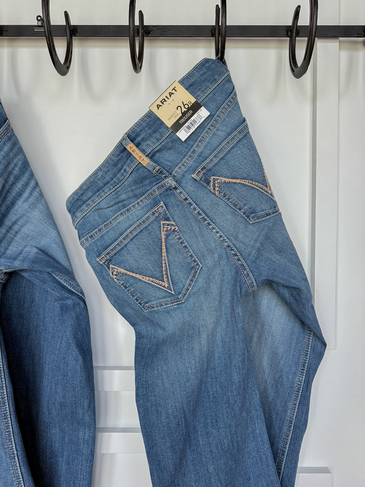 The Bowen Perfect Rise Trouser Jean by Savannah Sevens western life{&}style, in medium wash with a comfy no-gap waist, hangs on a black hook rack. The jeans have back pockets with tan stitching and a tagged barcode against a white wall.