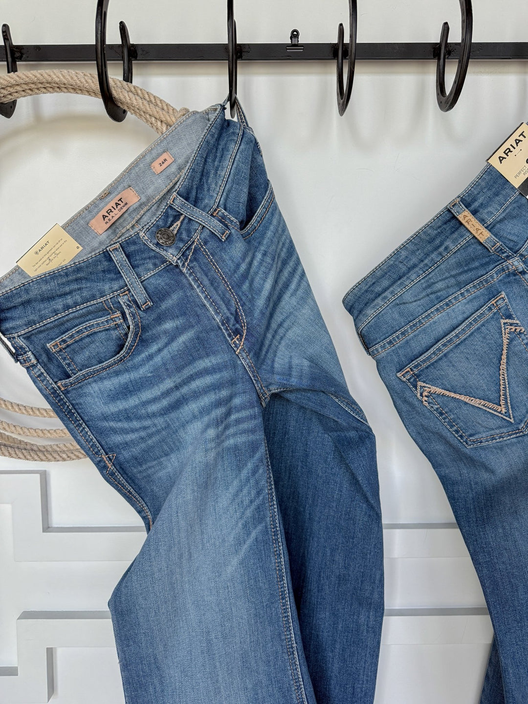 Two pairs of The Bowen Perfect Rise Trouser Jeans by Savannah Sevens western life{&}style hang on black hooks against a white wall. The denim features natural fading, stitching detail, tan waistband labels, and a coiled rope on the left hook.