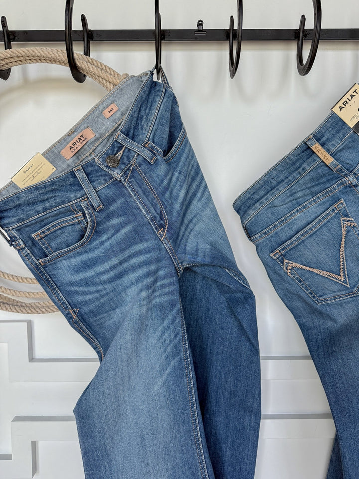 Two pairs of The Bowen Perfect Rise Trouser Jeans by Savannah Sevens western life{&}style hang on black hooks against a white wall. The denim features natural fading, stitching detail, tan waistband labels, and a coiled rope on the left hook.