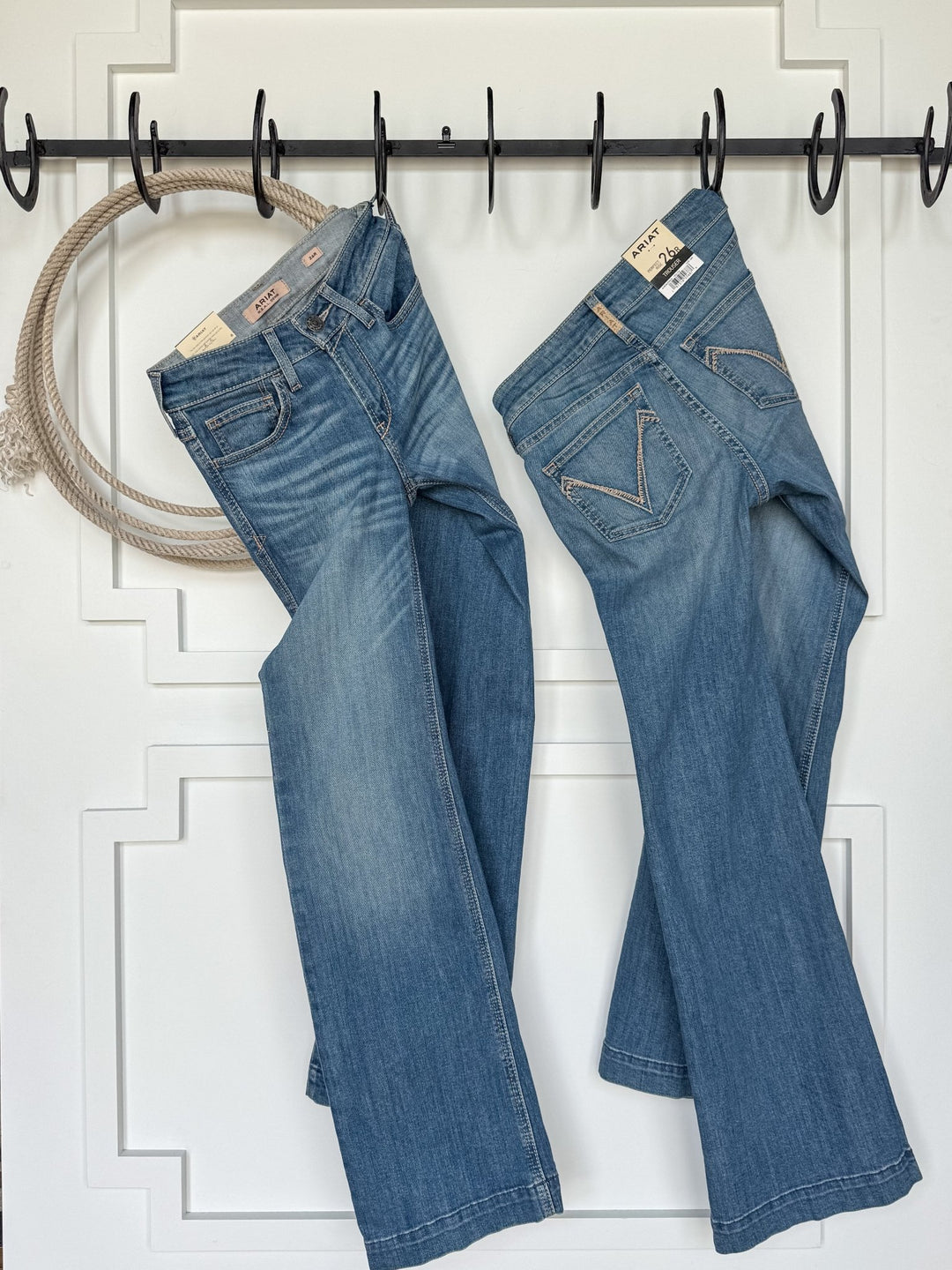 Two pairs of The Bowen Perfect Rise Trouser Jean by Savannah Sevens western life{&}style hang from black hooks on a white paneled wall with a coiled rope, set amid rustic wood and patterns—perfect for comfy no-gap waist denim lovers.