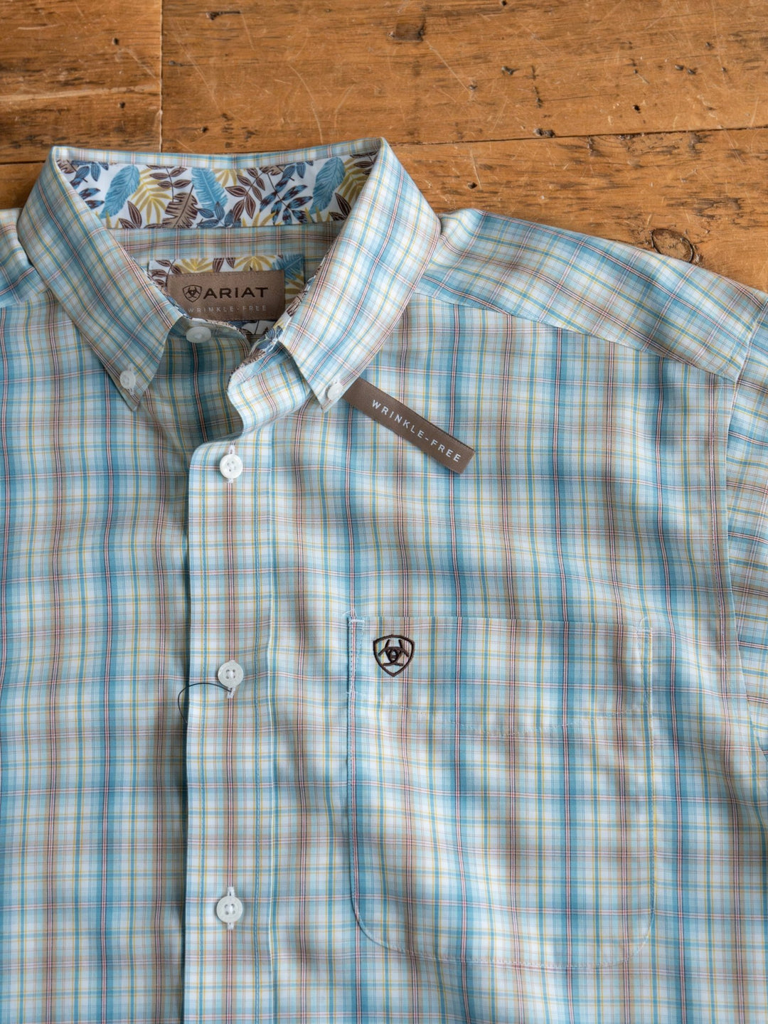 The Bowmont Men's Ariat Plaid Button Up from Savannah Sevens western life{&}style features blue, tan, and white plaid, a Wrinkle-Free chest tag, and a contrasting blue leaf inner collar. Displayed neatly folded on a wooden surface.