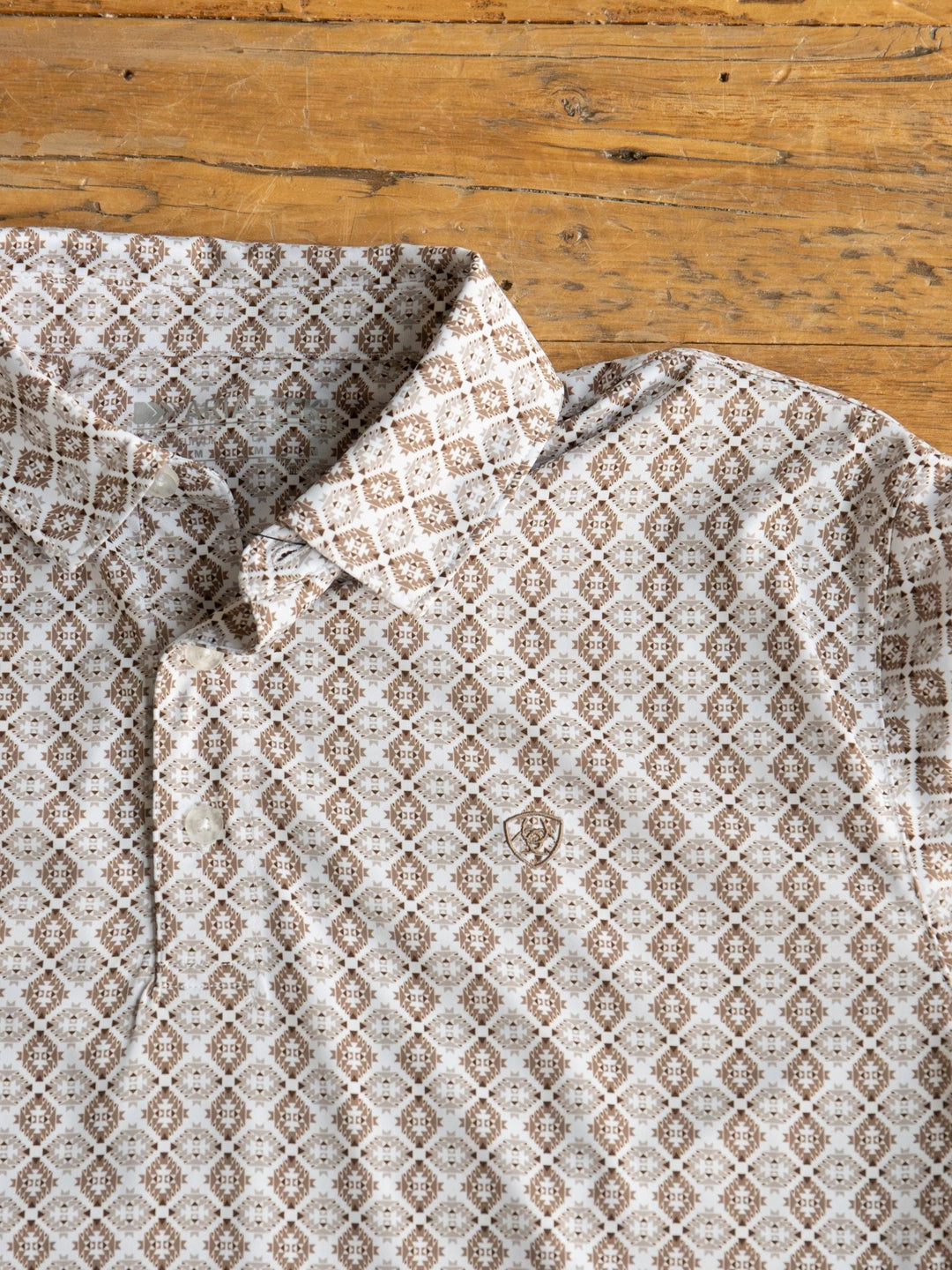 A close-up of the Bozeman Ariat Men's Southwest Polo by Savannah Sevens western life{&}style in beige and brown with a subtle chest crest and moisture-wicking fabric, laid flat on rustic wood.