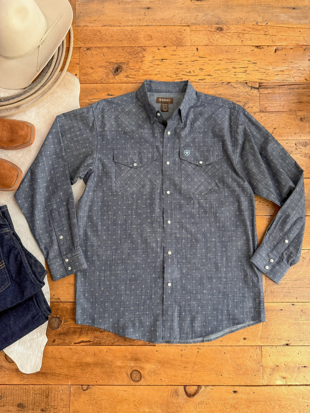 The Branford Ariat Men's Chambray Pearl Snap by Savannah Sevens western life{&}style is laid flat on a wooden floor, paired with a beige cowboy hat, brown boots, and dark blue jeans.