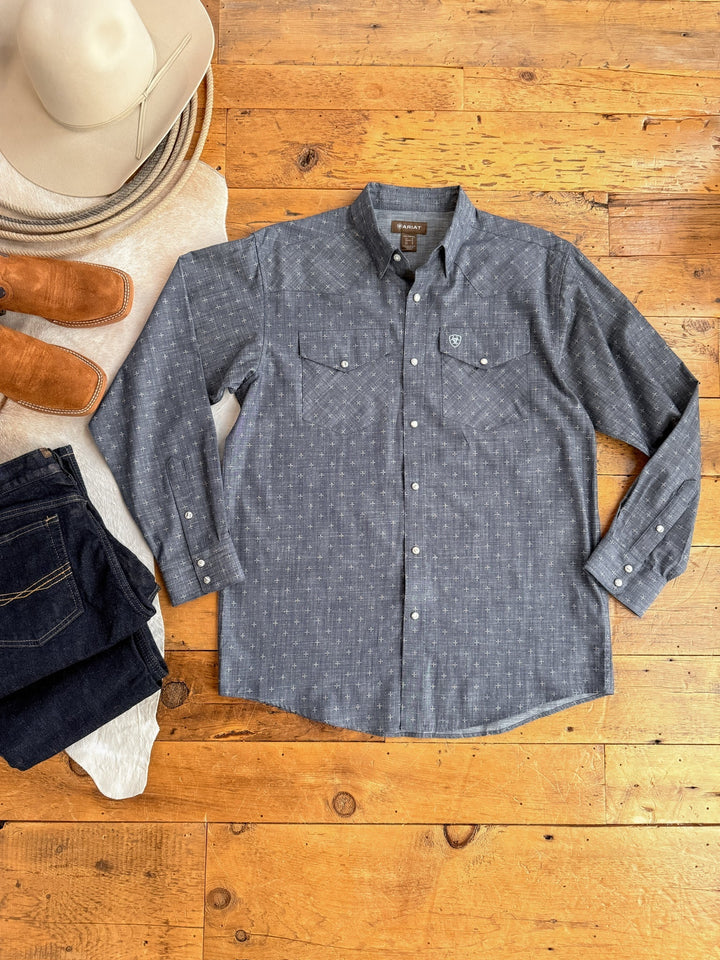 The Branford Ariat Men's Chambray Pearl Snap by Savannah Sevens western life{&}style is displayed on a wooden floor with dark jeans, brown cowboy boots, and a light-colored cowboy hat.