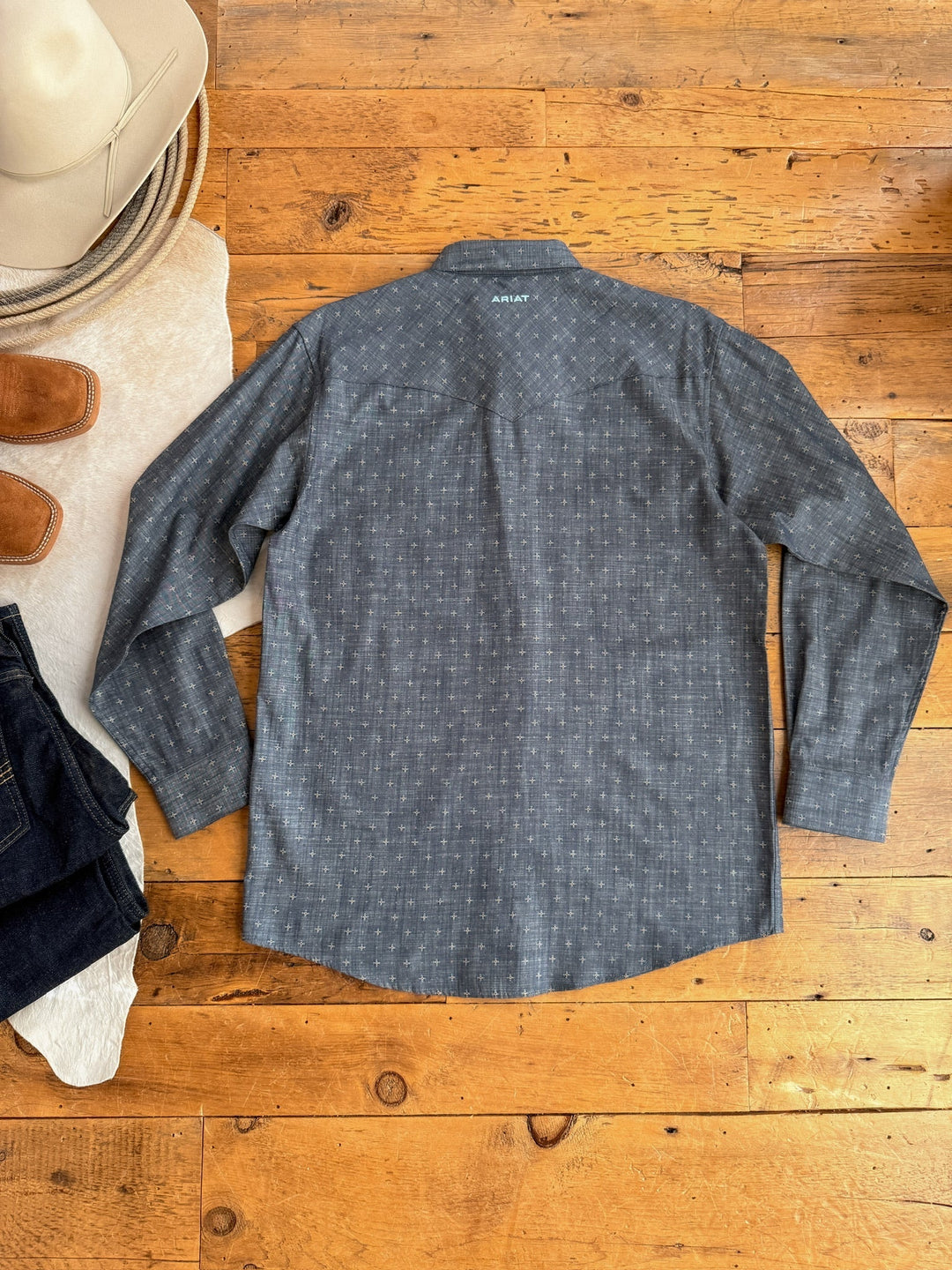 The Savannah Sevens western life{&}style Branford Ariat Men's Chambray Pearl Snap shirt in patterned gray is laid flat on a wooden floor with a tan cowboy hat, brown boots, part of a cowhide rug, and folded dark jeans.