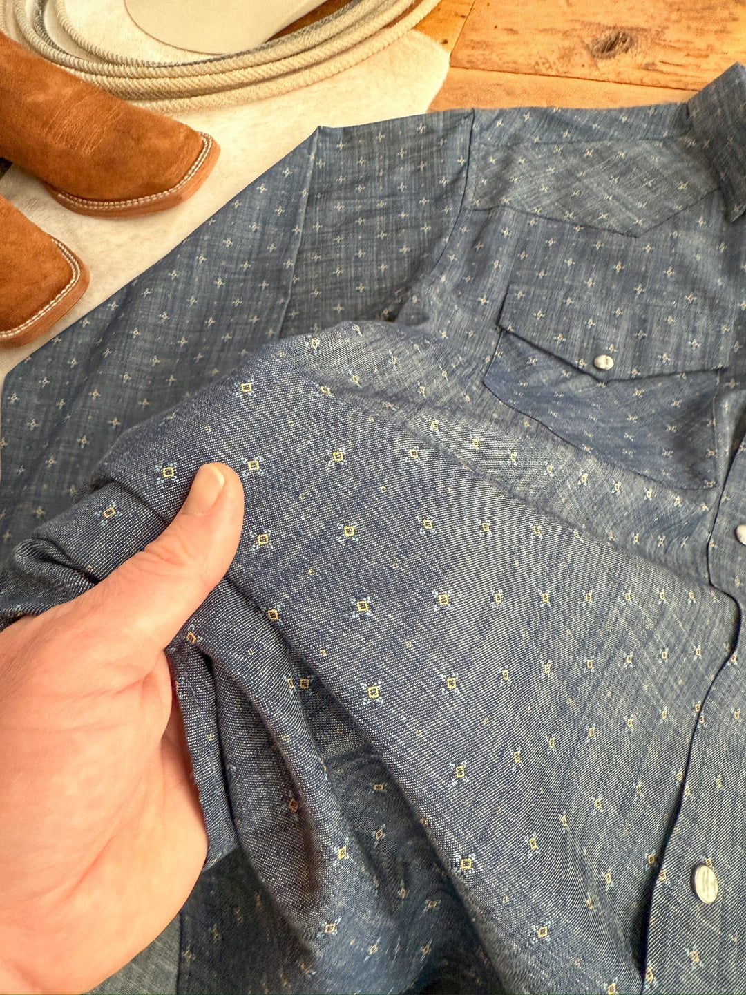 A hand holds the sleeve of the Branford Ariat Men's Chambray Pearl Snap shirt from Savannah Sevens western life{&}style, laid on wood with a tan boot and coiled rope in the background.