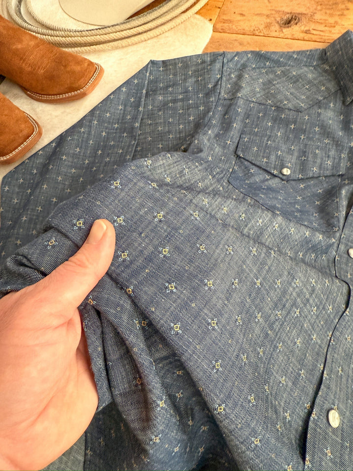 A hand holds the sleeve of the Branford Ariat Men's Chambray Pearl Snap shirt from Savannah Sevens western life{&}style, laid on wood with a tan boot and coiled rope in the background.