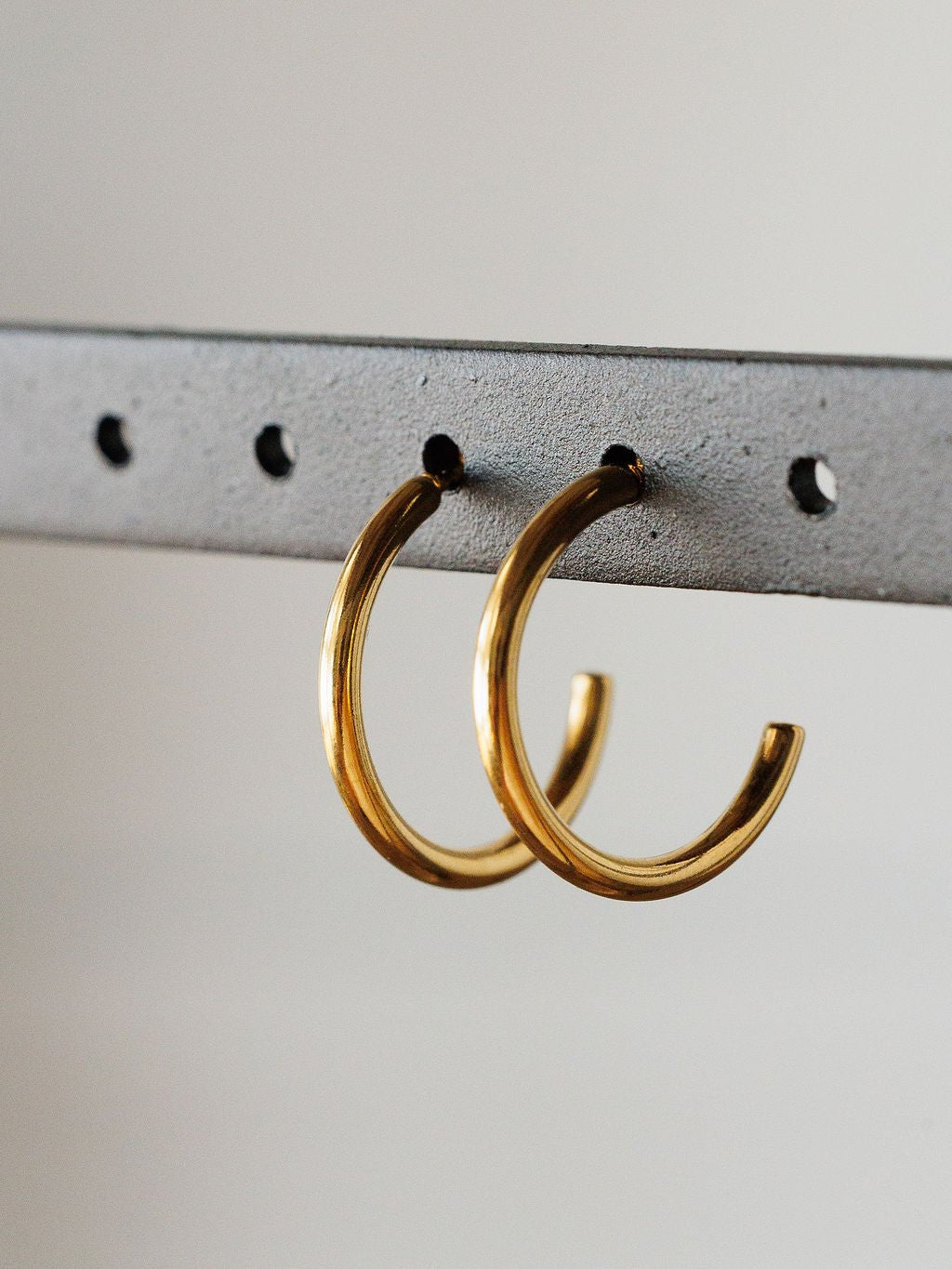 The Brazon Earrings by Savannah Sevens western life{&}style are gold mini hoops displayed on a gray metal bar with evenly spaced holes and a neutral background. These hypoallergenic earrings are perfect for everyday wear.