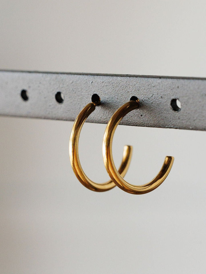 The Brazon Earrings by Savannah Sevens western life{&}style are gold mini hoops displayed on a gray metal bar with evenly spaced holes and a neutral background. These hypoallergenic earrings are perfect for everyday wear.