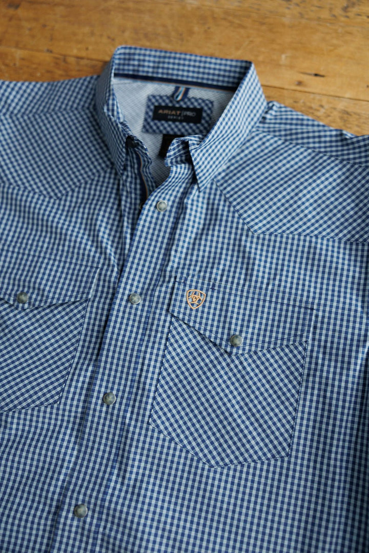 The Brentwood Ariat Men's Blue Plaid Pearl Snap shirt by Savannah Sevens western life{&}style features blue and white plaid, long sleeves, two chest pockets, pearl snap buttons, and an embroidered logo on one pocket, laid flat on wood.