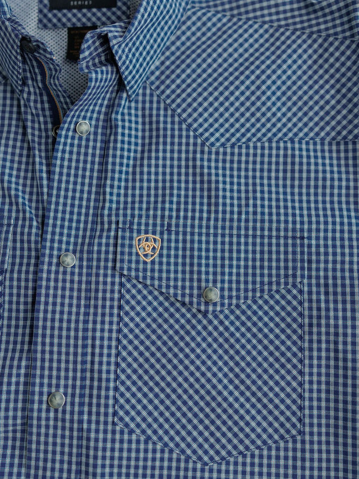 Close-up of the Brentwood Ariat Men's Blue Plaid Pearl Snap shirt from Savannah Sevens western life{&}style, featuring blue and white plaid fabric, pearl snap buttons, a chest pocket, and a small gold Ariat emblem on the pocket corner.